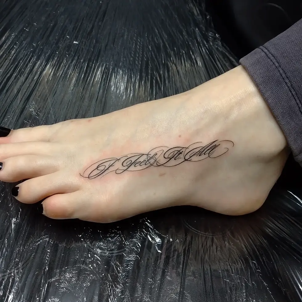 Close-up of a bare foot with black script tattoo reading I Feel It All, resting on shiny black plastic.