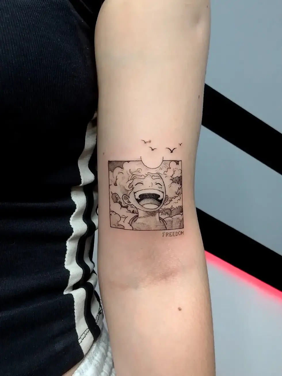 Forearm tattoo of a laughing anime face with birds and the word FREEDOM, on a person in a black shirt against a pale wall.