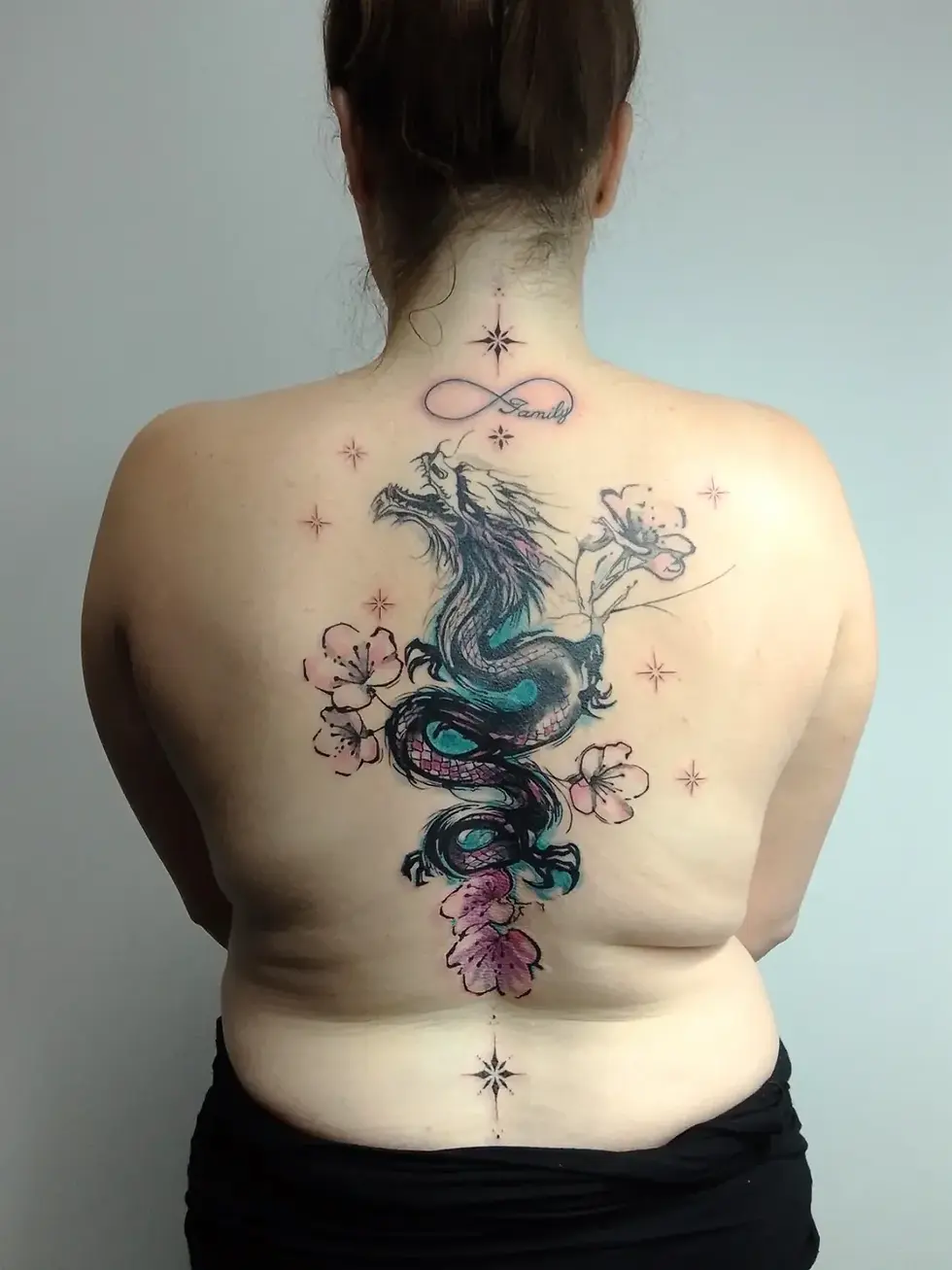 Back view of woman with large colorful dragon and cherry blossom tattoo, plus Family on neck, against plain gray wall.