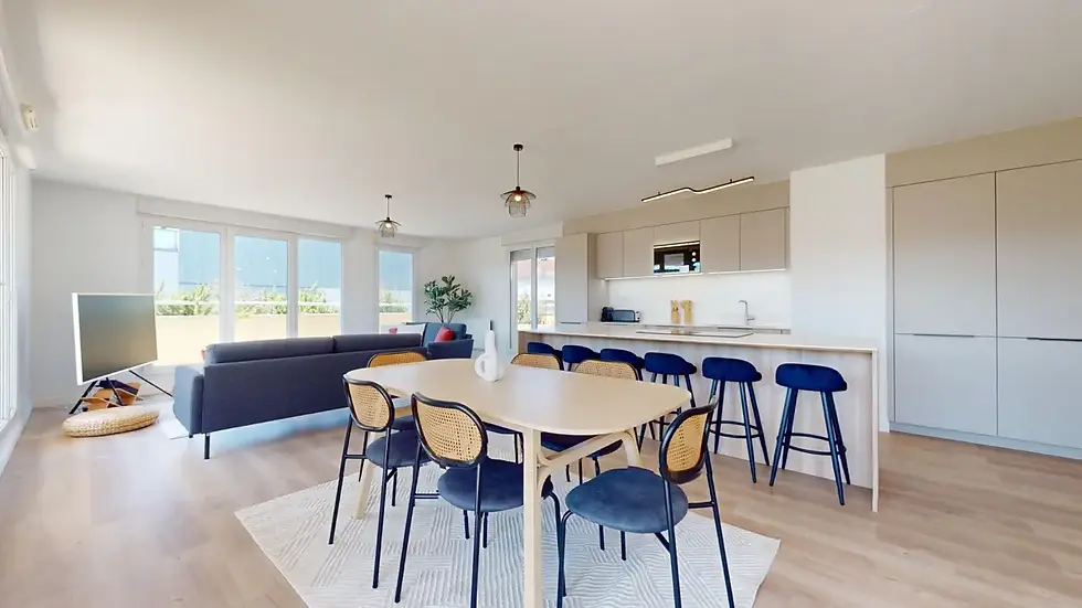 Pièce de vie lumineuse. Un coin repas comprend une table ovale en bois clair entourée de chaises noires et beiges avec des coussins bleus. Un salon confortable éclairé par de grandes fenêtres. Colocation Altitude à Annemasse