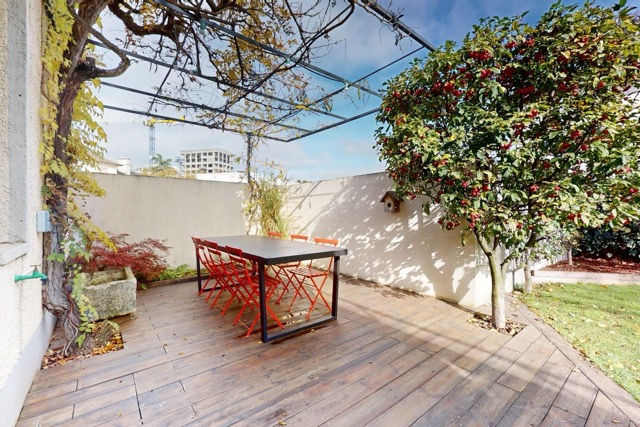 Aménager, meubler et décorer les extérieurs d'une colocation : terrasse en bois avec grande table noire, chaises orange vives et pergola végétalisée en jardin arboré.