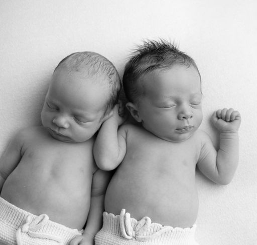 Newborn twin babies peacefully lying together, with their heads touching in a natural pose, captured in black and white.