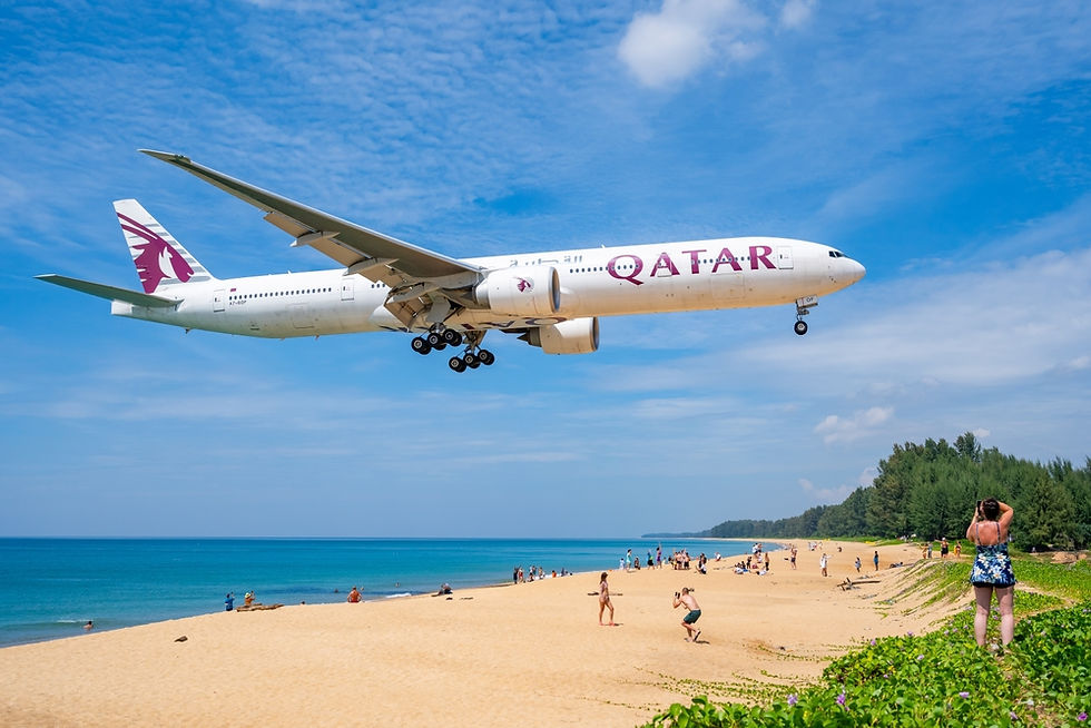 Airplane flying low over a beach with blue sky. "Qatar" written on plane. People on sand watch and take photos. Lush green vegetation nearby - หาดไม้ขาว วิวทะเลพร้อมกับเครื่องบินบนหาด