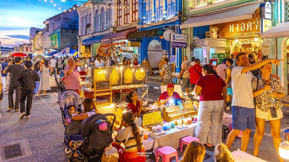 Bustling night market with colorful stalls, people browsing, vibrant lights, and a vendor selling candles. Signs for currency exchange. Phuket Cultural