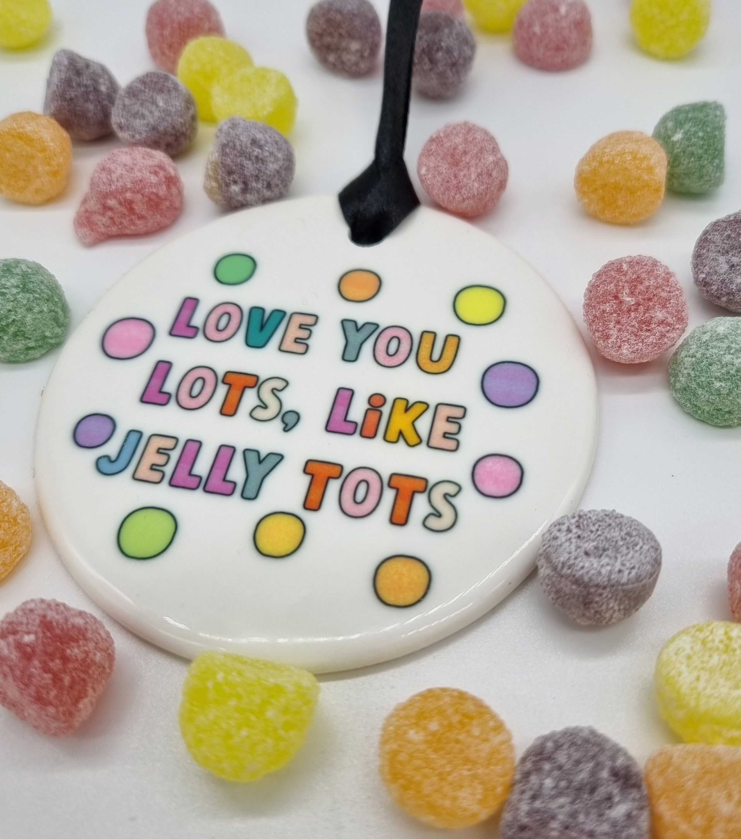 White round ceramic hanging decoration on a black ribbon. Colourful bubble text that says "Love you lots like Jelly Tots'