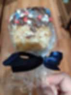 Dipped Rice Krispie treat with a layer of chocolate near the top and red, white, and blue sprinkles shaped like stars. The treat is on a stick and wrapped in a clear plastic bag tied up with a blue ribbon, being held by a white hand.