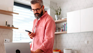 Homem barbudo de camisa rosa usa celular com aplicativo do Albert ao lado de laptop na cozinha clara, com expressão concentrada.