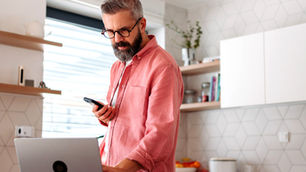 Homem barbudo de camisa rosa usa celular com aplicativo do Albert ao lado de laptop na cozinha clara, com expressão concentrada.