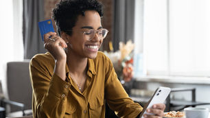 Mulher sorridente com óculos segura cartão azul e celular à mesa, em ambiente claro, com xícara ao lado.