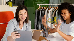 Duas mulheres sorrindo em escritório de moda, uma usa tablet e a outra faz gesto de ok; arara de roupas ao fundo.