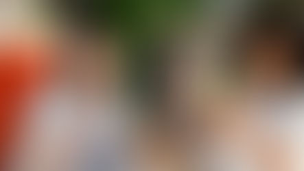 Duas mulheres sorrindo em escritório de moda, uma usa tablet e a outra faz gesto de ok; arara de roupas ao fundo.