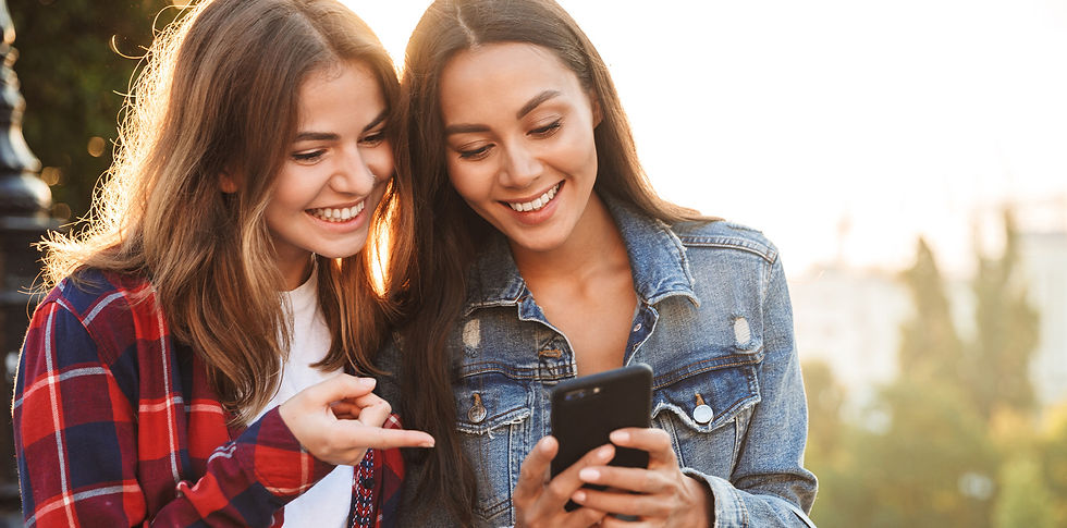 Duas jovens sorrindo olham um celular ao ar livre, com luz dourada ao fundo; uma aponta para a tela.