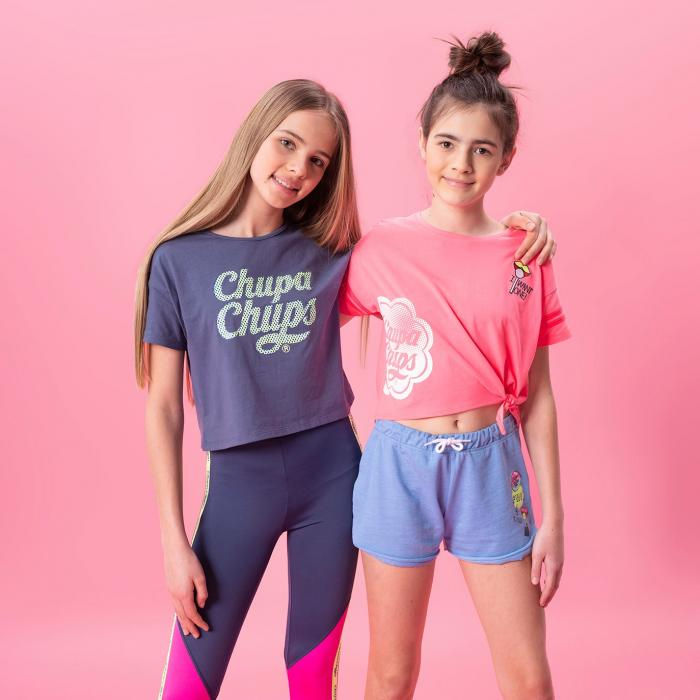 Two young girls hugging each other in front of a pink wall. One girl is wearing a purple t-shirt and purple pants and the other girl is wearing blue shorts and a pink top.