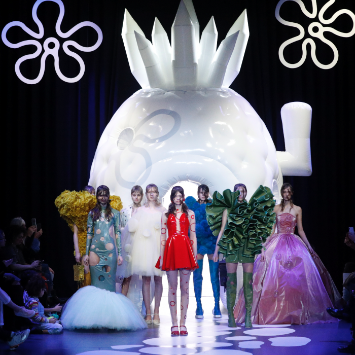 A group of models standing in the middle of a runway, wearing colourful outfits. Behind them is a large, white structure.