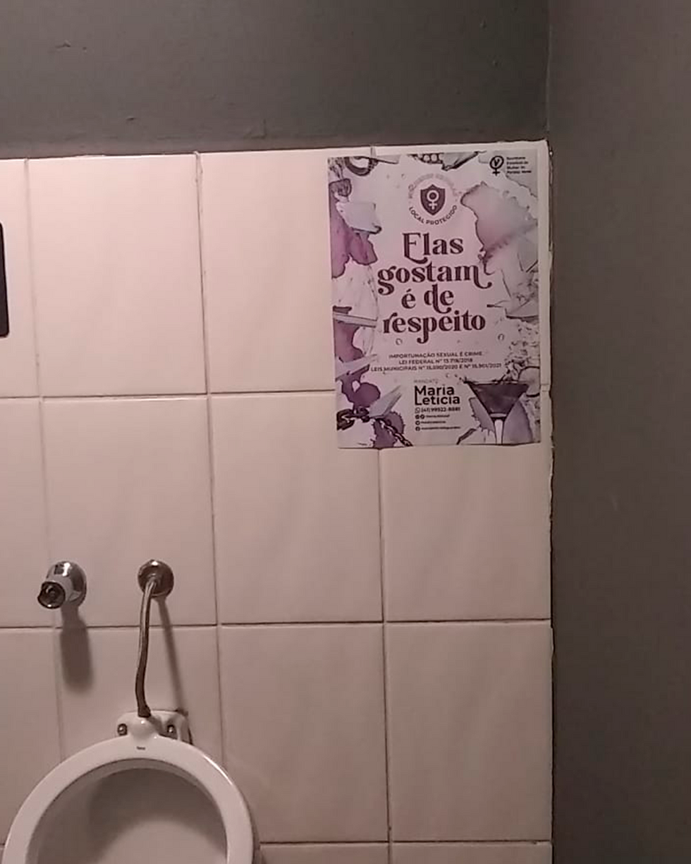 Cartaz colado em uma parede ao lado de um mictório masculino com a frase "Elas gostam de respeito"