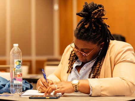 Woman with locs in a blazer that is writing with a blue pen in a notebook. She's focused and has a water bottle sitting nearby.
