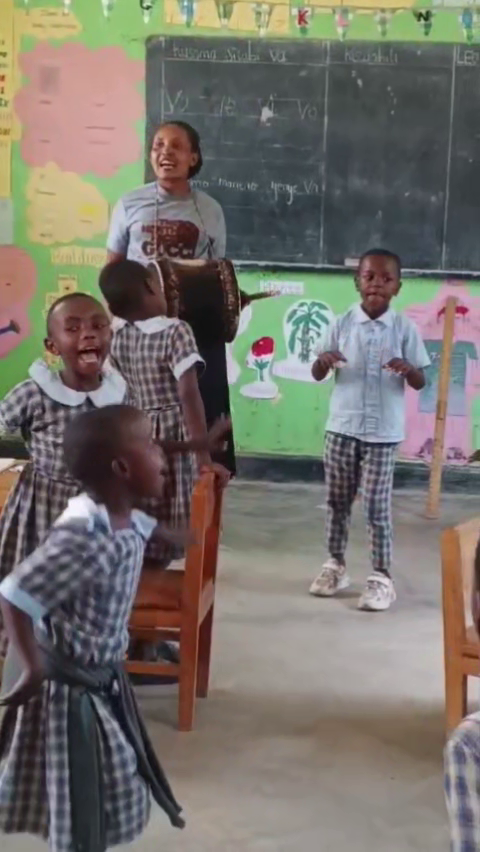 Photo verticale en classe en Afrique, des enfants en uniforme qui dansent et une adulte qui joue du tambour, ambiance joyeuse et éducative, couleurs vives, format portrait.