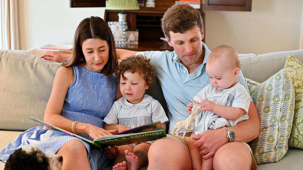 Family of four on a couch reading a picture book, with two toddlers and a baby; cozy living room, calm and affectionate mood.