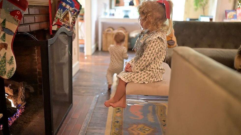 Toddler in festive dress sits by a fireplace with Christmas stockings, while another child stands in a cozy living room.