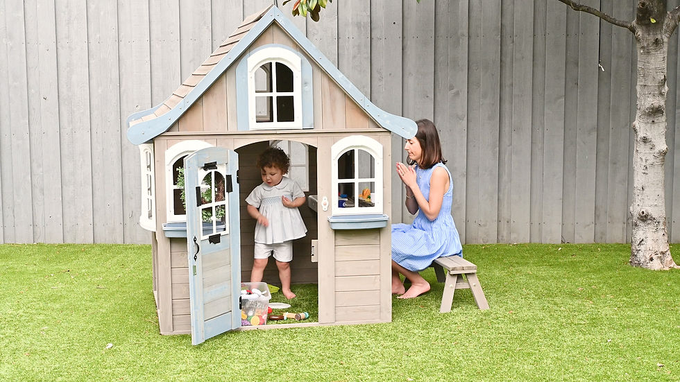 Woman kneels by a wooden playhouse as a toddler stands in the doorway, in a sunny backyard with grass and a gray fence.