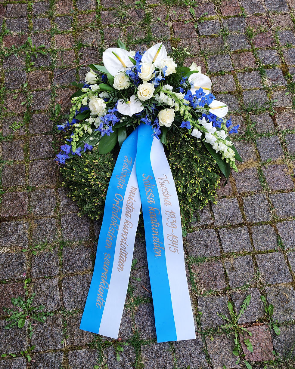 Seppele sisältää sini-valkoisen seppelenauhan, johon teksti käsin tekstattuna.
