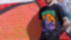 A bearded man with tattoos leaning against a brick wall with a Denton Halloween shirt on