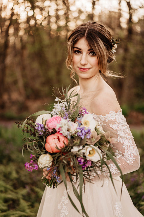 Wearing a bohemian style wedding dress that sits off her shoulders, but has long sleeves and is made of delicate lace, this bride is holding a large, natural style bouquet of flowers which includes peonies and sweet Williams. A soft flowing hairstyle, with face framing pieces compliments the look. 