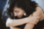 A close-up photograph of a woman with dark hair. Her arms are crossed and she is holding onto herself, appearing sad or in discomfort.