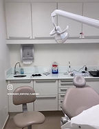Foto de um consultório dentario, com uma cadeira de dentista, uma cadeira para o dentista e armários brancos.