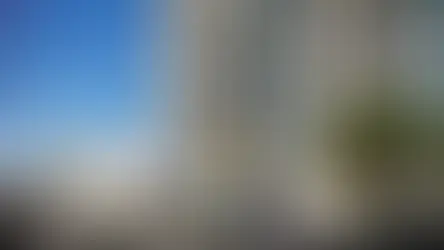 Prédio residencial moderno com varandas, pavilhão de vidro na calçada, árvores e carros sob céu azul ensolarado.