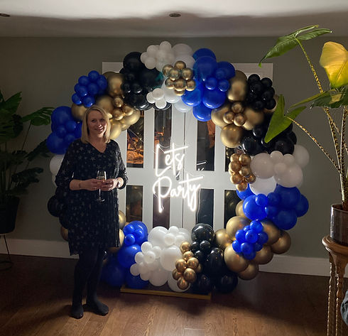 Photo of owner standing by a balloon arch of blue, white and gold balloons that says Lets Party in neon lights