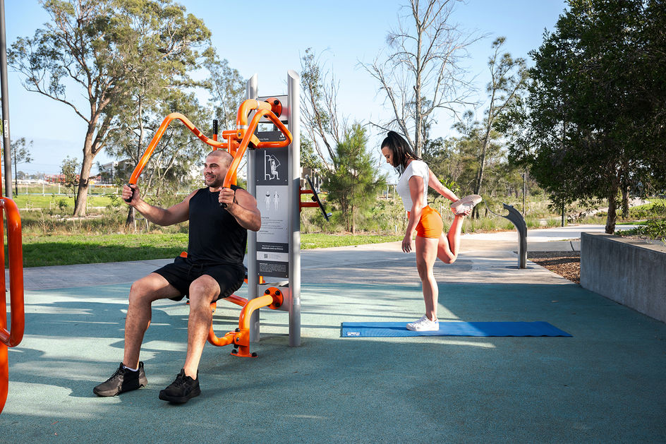 Man using outdoor gym equipment while a woman stretches on a mat in a public park fitness area