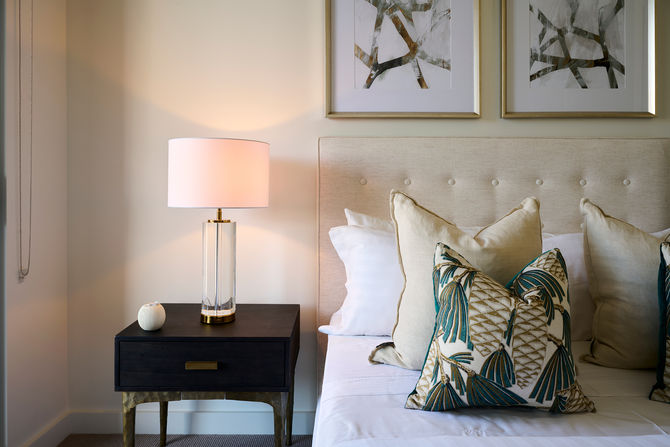 Close-up of a stylish apartment bedroom featuring a neutral upholstered headboard, layered decorative cushions, and a modern bedside table with a sculptural lamp, highlighting refined interior styling, luxury residential design, and comfortable contemporary living.