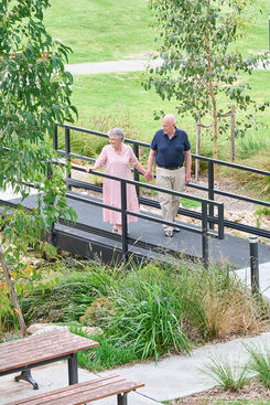 An older couple walking hand in hand across a garden pathway surrounded by greenery. Two Carrington Care residents walking together along an accessible garden walkway in a landscaped outdoor setting. Carrington Care residents enjoying a walk together along a safe, accessible pathway through landscaped gardens. Two Carrington Care residents walking hand in hand along an accessible garden path, highlighting connection, independence, and wellbeing in an outdoor setting. Residents walking together through landscaped gardens.