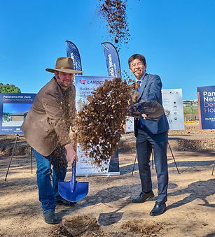 Two men in business attire and a hat shovel and toss dirt into the air during a groundbreaking ceremony at a construction sit
