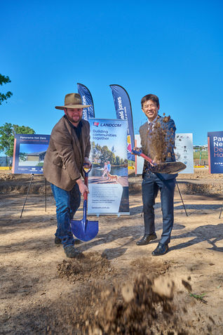 Two men in business attire perform a ceremonial sod turning at a Landcom development site, tossing soil with shovels in front of project signage under a clear blue sky.