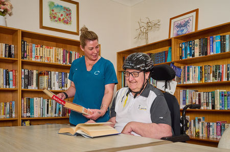 A care worker and an older man reading together at a table in a library setting. A Carrington Care team member sharing a book with a resident in a library space, surrounded by bookshelves. A Carrington Care support worker reading with a resident in a library, highlighting connection, learning, and social engagement. A Carrington Care staff member engaging with a resident as they read together in a warm, book-filled library space, reflecting personalised and supportive care. Shared reading time in an aged care library.