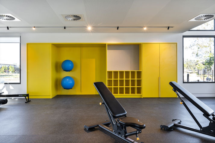 Modern fitness centre at Stockland Halcyon Gables featuring adjustable weight benches, exercise equipment, bright yellow storage cabinetry with stability balls, rubber flooring, and large windows providing natural light in a contemporary resident gym.
