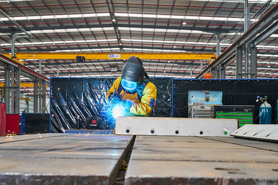 Welder wearing protective gear welding a metal component inside a factory. A welder in protective equipment welding a steel component on the factory floor, with sparks visible. Skilled welder carrying out metal fabrication work in an industrial workshop, with sparks flying during the welding process. A welder in full protective gear welding a steel component inside a large industrial workshop, capturing precision, safety, and craftsmanship in manufacturing. Metal fabrication in progress as a welder joins steel components inside an industrial facility.