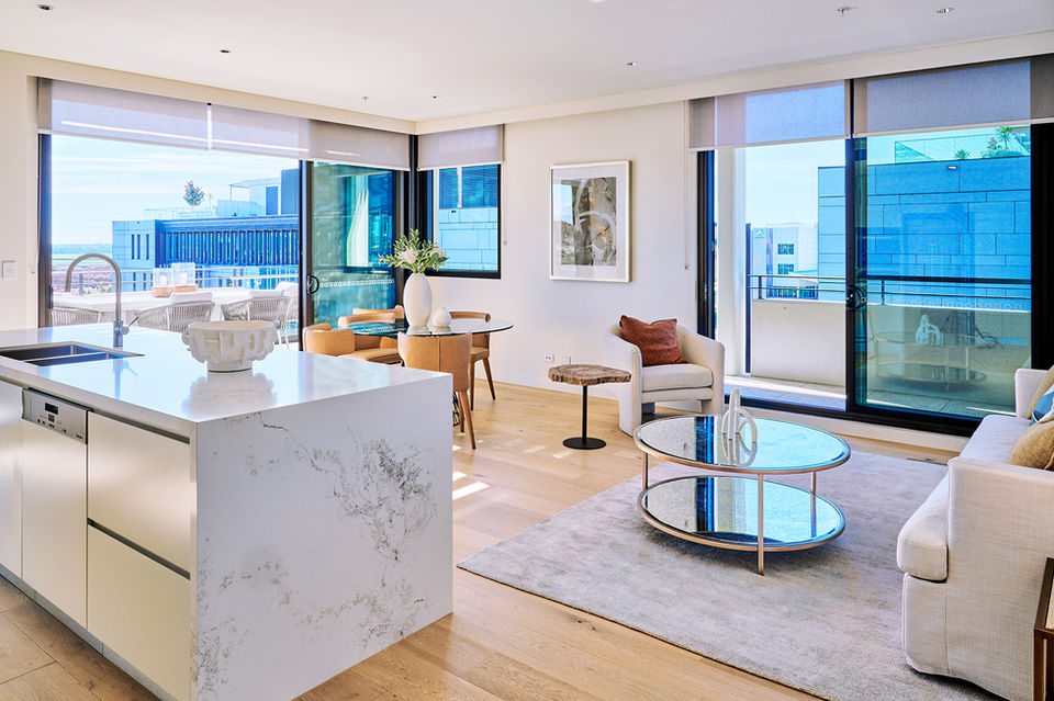 Light-filled open-plan apartment interior with a modern kitchen island, dining area, and living space, featuring floor-to-ceiling windows and sliding doors to a private balcony, showcasing contemporary design, premium finishes, and seamless indoor–outdoor apartment living.