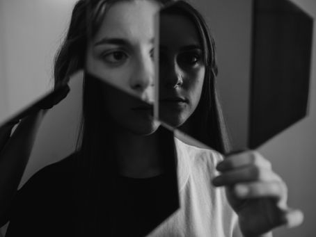 A woman holds fragmented mirror pieces, reflecting different parts of her face in a striking monochrome portrait.