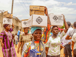 Women carry aid boxes on heads on a dusty street; boxes read Lagos Food Bank Initiative.
