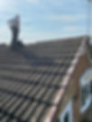 A worker stands on a sloped, tiled roof, surveying the area under a bright blue sky. Dust and debris are visible on the tiles