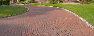 A curved brick driveway with a herringbone pattern, bordered by neatly trimmed grass and stone edging