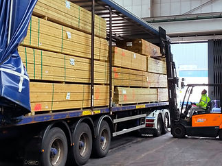 A forklift operator unloads stacked wooden planks from a truck trailer inside a warehouse. Brightly lit industrial setting
