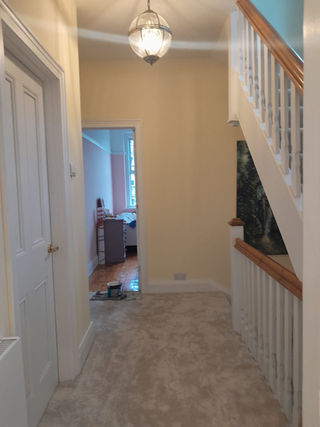 A well-lit hallway showcasing beige walls, a decorative light fixture, plush carpeting, and an open staircase leading upwards