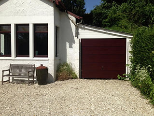 A white house with a brown garage door to the right, surrounded by greenery