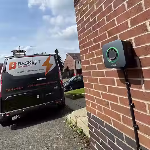 A black van with "Baskett Electrical Ltd" on it is parked near a brick wall with an electric vehicle charger