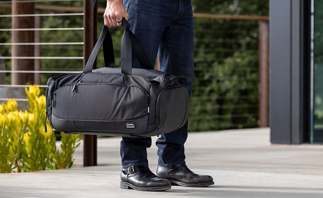 Person holding a large black duffel bag, standing outdoors near yellow flowers