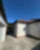 A sunlit corner of a house with a red-tiled roof, white walls, and a concrete area beside it under a clear blue sky